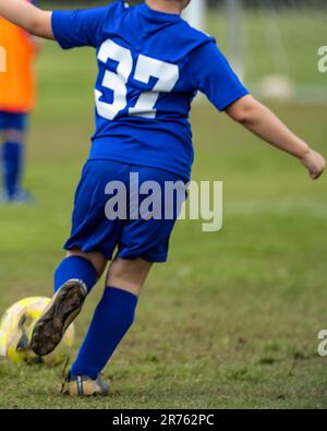 Un jeune garçon portant un maillot bleu donne un coup de pied à une balle de football sur un terrain de football Banque D'Images