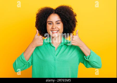 Joyeuse excitée curly cheveux brésilienne ou afro-américaine élégante positive femme, en chemise verte, regarde avec joie l'appareil photo et le sourire, montre le pouce vers le haut geste, se tient sur un fond jaune isolé Banque D'Images