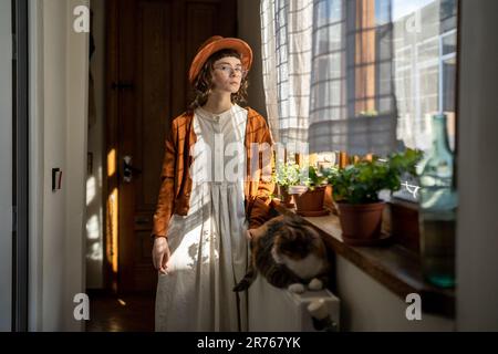 Pensive hipster fille propriétaire d'animal de compagnie debout près de chat moelleux allongé sur la batterie de chauffage à la maison Banque D'Images