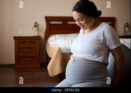 Bonne femme enceinte adulte, femme enceinte expectative assise sur le sol, souriant regardant son grand ventre à la fin de la grossesse Banque D'Images