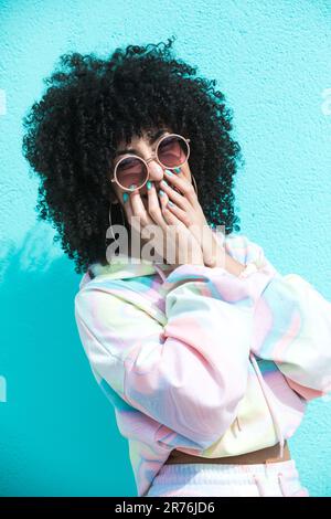 Bonne jeune femme hispanique avec une coiffure afro dans des lunettes de soleil couvrant la bouche avec les mains tout en regardant l'appareil photo et en riant Banque D'Images