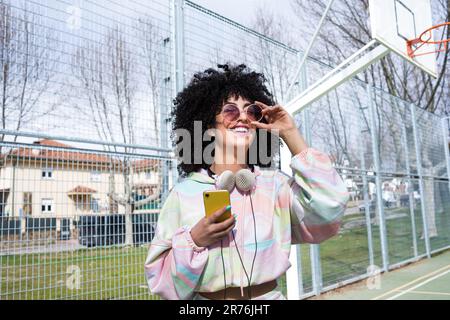 Positive jeune hispanique féminine avec Afro coiffure lunettes de soleil casque et smartphone dans la main souriant tout en ajustant les lunettes de soleil dans le terrain de sport Banque D'Images