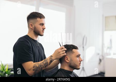 Vue latérale d'un jeune barbier sérieux avec tatouages coupant les cheveux de l'homme avec des ciseaux sur un arrière-plan flou dans un salon de coiffure Banque D'Images