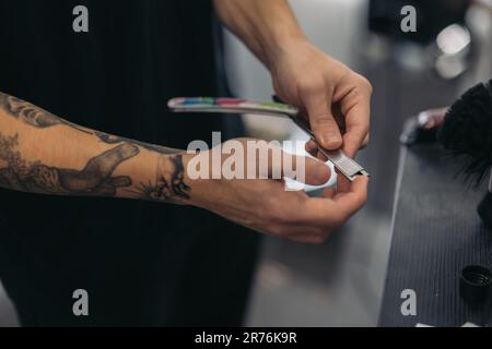 barbier anonyme court grand angle avec tatouages changeant le rasoir pour couper la barbe sur un arrière-plan flou Banque D'Images