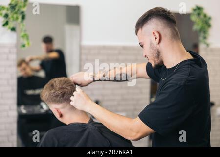 Vue latérale d'un jeune barbier avec tatouages coupant les cheveux d'un homme méconnaissable avec des tondeuses dans le salon de coiffure sur un arrière-plan flou Banque D'Images