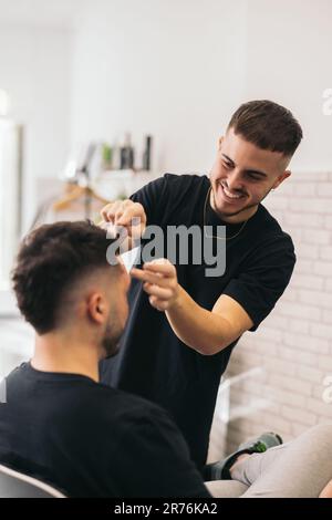 Vue de face d'un jeune barbier peignant un homme méconnaissable dans un salon de coiffure sur un arrière-plan flou Banque D'Images