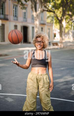 Joueuse de basket-ball afro-américaine en vêtements et lunettes ethniques debout dans la rue avec terrain de sport et regardant la caméra tout en lançant le ballon Banque D'Images