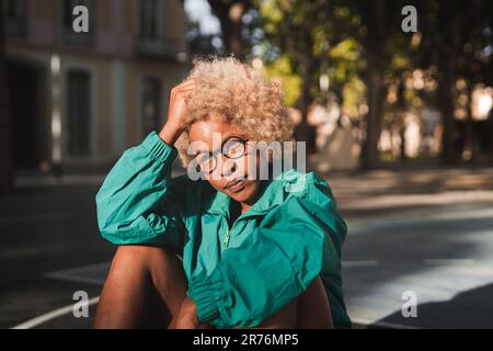 Bonne jeune athlète afro-américaine en vêtements de sport avec lunettes assises sur le terrain de basket-ball au coucher du soleil Banque D'Images