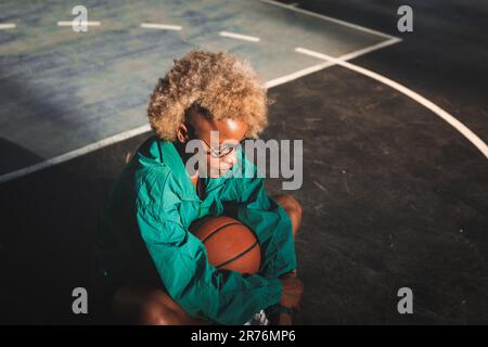 D'en haut, jeune femme afro-américaine pensive de basket-ball dans les cheveux blonds bouclés avec veste confortable et ballon assis sur le terrain de sport en soirée Banque D'Images