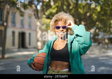 Jeune femme positive dans une tenue tendance et des lunettes de soleil souriant et regardant l'appareil-photo tout en se tenant debout avec le basket-ball sur la rue de la ville le jour ensoleillé Banque D'Images