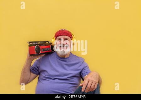 Portrait d'un homme senior à barbe gai dans une tenue décontractée et un casque tout en tenant un lecteur de cassette rétro et assis sur fond jaune Banque D'Images