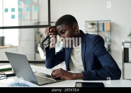 Jeune homme d'affaires afro-américain tendu assis sur le lieu de travail devant un ordinateur portable au bureau et regardant l'écran avec les données tout en les analysant Banque D'Images