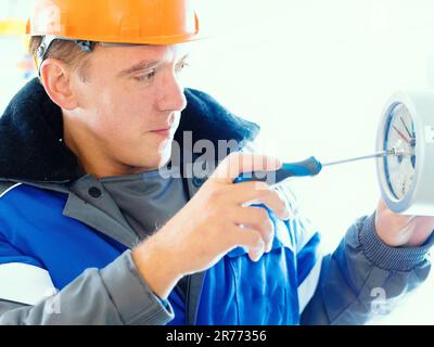 Un électricien ou un monteur d'instrumentation muni d'un tournevis à la main règle le fonctionnement d'un manomètre dans une installation industrielle de gaz intérieure. Travailleur caucasien dans un casque de sécurité orange et un pelage de pois d'hiver au travail. Portrait en gros plan. Banque D'Images