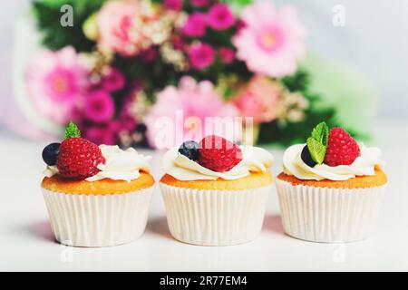 3 cupcake à la crème au beurre et aux baies fraîches, bouquet de fleurs roses sur fond Banque D'Images