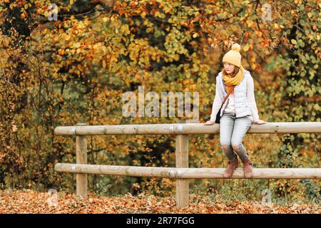 Portrait d'automne d'une adorable jeune fille de 10-11 ans, portant une veste en duvet grise, un chapeau et une écharpe jaunes, assis sur la clôture Banque D'Images