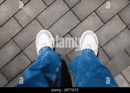 Jambes pour femmes en jeans et baskets blanches modernes sur carreaux gris, espace pour le texte, vue du dessus, point de vue personnel Banque D'Images