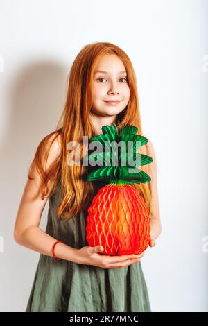 Photo de studio de la jeune fille adorable de la préadolescence à cheveux rouges, portant une robe kaki, jouant avec de l'ananas en papier Banque D'Images