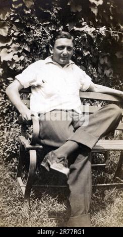 Carte postale photographique vintage d'un jeune homme assis à l'extérieur sur un banc de parc, vers 1925. Banque D'Images