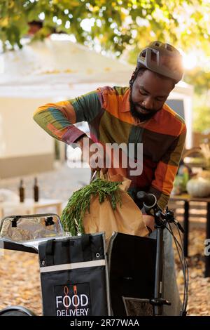 Jeune afro-américain mettant bio produits bio bio dans sac thermique, prenant la commande du marché agricole et de vélo d'équitation. Livraison de fruits et légumes cultivés localement à des clients de la ferme. Banque D'Images