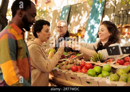 Divers couple homme et femme achetant des produits naturels frais sur le marché des agriculteurs, regardant les produits bio. Un fournisseur local souriant, debout derrière un stand de fruits et légumes, vend des aliments sains. Banque D'Images