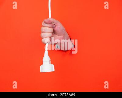 La main d'un homme tient un avec câble d'alimentation à travers du papier orange déchiré. Passez le papier orange déchiré. Banque D'Images