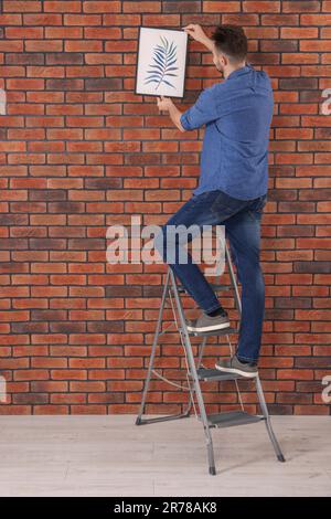 Homme sur un escabeau accrochant une photo près du mur de briques rouges Banque D'Images