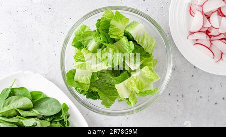 Salade de printemps dans un bol en verre. La laitue romaine, les jeunes épinards et les tranches de radis frais sont en gros plan sur la table de cuisine, à plat avec espace pour les copies Banque D'Images