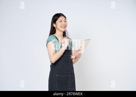 Jeune femme portant un tablier de cuisine tenant bloc-notes livre de cuisine recette et stylo isolé sur fond blanc. Banque D'Images