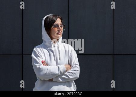 Portrait femme brune mûre avec capuche sur la tête, porte un sweat à capuche blanc et des lunettes de soleil avec bras croisés, mur de fond sombre. Sportive confiant, 40 ans Banque D'Images