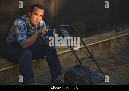 Portrait d'un jardinier regardant l'appareil photo, assis près d'une tondeuse avec une tasse de café entre les mains, reposant au coucher du soleil Banque D'Images