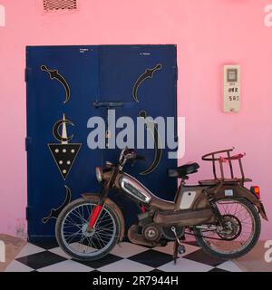 Mirleft, Maroc - Motobécane vintage moto s'appuie sur une double porte marocaine bleue avec des symboles. Véhicule stationné sur carreaux à carreaux noirs et blancs. Banque D'Images