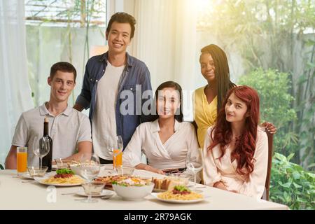 Cinq amis interculturels joyeux assis et debout près d'une table servie avec une variété de plats maison appétissants préparés pour la fête Banque D'Images