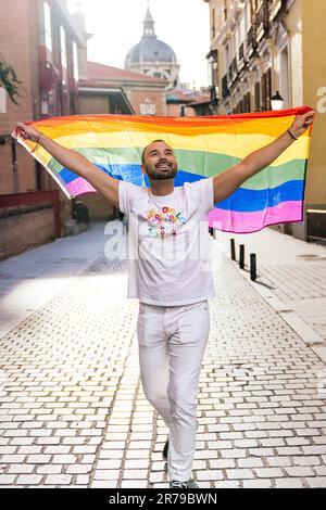 Homme à barbe, avec vue sur le ciel, portant un drapeau LGBT ouvert, au milieu d'une rue ensoleillée au crépuscule. Banque D'Images