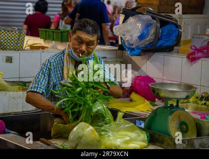 2023- 24 mars- MALAISIE, PENANG, GEORGETOWN -vendeur de légumes sur le marché avec une cigarette allumée dans sa bouche coupant des légumes Banque D'Images