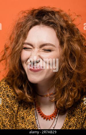 Jeune femme heureuse aux cheveux rouges avec maquillage naturel portant des colliers et un chemisier jaune avec imprimé abstrait isolé sur orange, élégante tenue décontractée a Banque D'Images