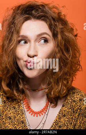 Portrait d'une jeune femme à cheveux rouges en blouse jaune et colliers qui se vissent sur les lèvres et regardent loin tout en se tenant isolé sur l'extérieur orange, élégant et décontracté Banque D'Images