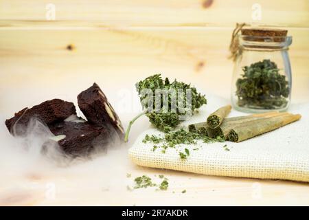 Pré-rouler les joints de marijuana et les brownies de gâteau avec des boutons de cannabis dans un pot en verre transparent qui pose sur la toile à biscuits Banque D'Images