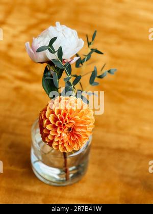 Composition florale de pivoine rose, de dahlia orange et de brindilles avec feuilles dans un vase en verre. Belle vie de fleurs et pot transparent sur un bois Banque D'Images