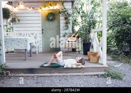 Une petite fille se trouve sur des oreillers sur la terrasse de la maison rustique. Un adorable enfant rêveur a mis ses mains sous la tête et regarde le ciel. Intérieur rétro avec table en bois Banque D'Images