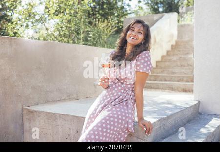 Belle jeune femme de race mixte en robe rose souriant et tenant un verre de champagne. Jolie fille s'assoit sur un parapet en pierre et boit un délicieux vin Banque D'Images