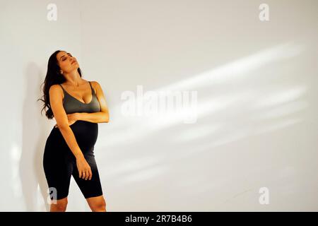 Belle femme enceinte en vêtements sombres se tient latéralement contre le mur blanc. Une charmante brunette attend bébé qu'elle mette sa main sur son ventre et tient le pic Banque D'Images