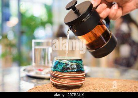 La main de femme verse le thé d'une presse française dans une belle tasse faite à la main sur une table dans un café, mise au point sélective, arrière-plan flou. Style de vie, réunion Banque D'Images