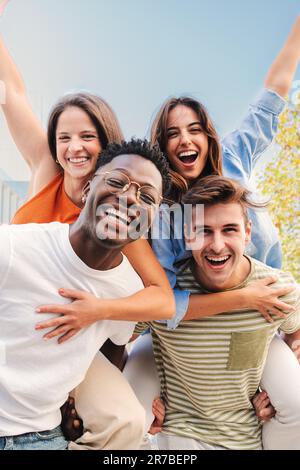 Portrait vertical de deux jeunes amis multiraciaux offrant une promenade en porcgyback à ses amies. Un groupe de gens heureux qui s'amusent et sourient. Des hommes portant des femmes sur ses épaules regardant l'appareil photo. Photo de haute qualité Banque D'Images