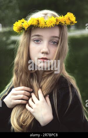 Une fille sérieuse et mystérieuse dans une couronne de pissenlits. Une fille aux cheveux longs et épais est triste au printemps dans la rue. Banque D'Images