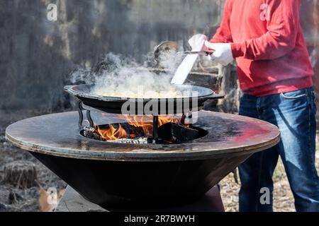 Les légumes sont rôtis sur un foyer rond. Gril rond en forme de bol avec un feu à l'intérieur. L'homme prépare le foyer pour le barbecue. Le bois de chauffage est en feu Banque D'Images