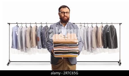 Homme barbu tenant une pile de vêtements pliés devant des porte-vêtements isolés sur un fond blanc Banque D'Images