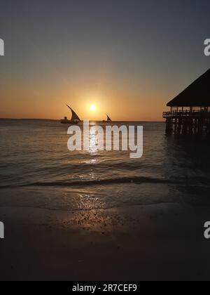 Kendwa, Île de Zanzibar, Tanzanie dhow bateaux voile et un quai de bungalow en bois de chaume contre le soleil couchant et ciel nuageux. Banque D'Images