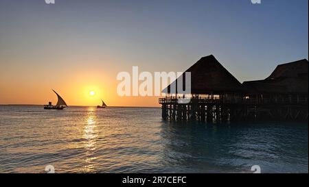 Kendwa, Île de Zanzibar, Tanzanie dhow bateaux voile et un quai de bungalow en bois de chaume contre le soleil couchant et ciel nuageux. Banque D'Images