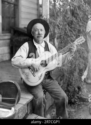 États-Unis: c. 1925 Portrait d'un homme âgé jouant une guitare sur son porche. Banque D'Images