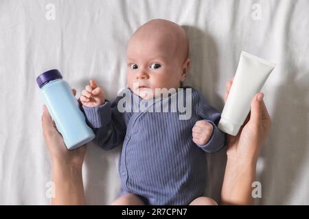 Mère tenant de la crème pour le corps et de la poudre à dépoussiérer près de mignon petit bébé sur le lit, vue du dessus Banque D'Images